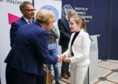 Dziewczynka ubrana w biały garnitur odbiera gratulacje od Małopolskiego Kuratora Oświaty. Dziewczynka stoi po prawej stronie zdjęcia.