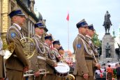 Żołnierze Wojska Polskiego stojący pod pomnikiem podczas występu artystycznego. W dłoniach trzymają instrumenty.