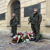 Żołnierze stojący przy tablicy pamiątkowej w pełnym umundurowaniu, na środku widzimy złożone wieńce przez różne organizacje, w tym Kuratorium Oświaty