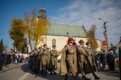 Przemarsz kolumny wojskowej. Na przodzie trzech mężczyzn z biało-czerwonym sztandarem. W tle kościół.