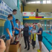 Zawodnicy stoją na podium w hali basenowej, Artur Pasek dekoruje laureata III miejsca, obok stoi trenerka z tacą z medalami.