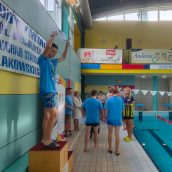 Zwycięzca na podium w hali basenowej, w podniesionych rękach trzyma dyplom.