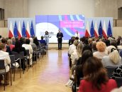 Ministra edukacji Barbara Nowacka oraz inni oficjele w trakcie swojego wystąpienia. Na bliższym planie słuchająca publiczność, po prawej flagi Polski oraz Unii Europejskiej.