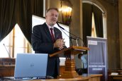 I Małopolski Wicekurator Oświaty stojący na scenie i przemawiający do zgromadzonych osób w trakcie konferencji, za nim baner reklamowy, przed nim laptop.