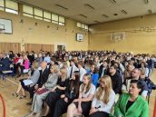 Widok ogólny sali gimnastycznej, gdzie odbywało się zakończenie roku, na zdjęciu uczniowie, absolwenci oraz nauczyciele siedzący na krzesłach.
