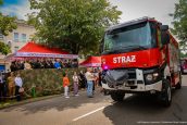 Czerwony wóz strażacki jadący ulicą w trakcie trwania zakończenia roku, obok niego obserwujący go widzowie, zwróceni w stronę samochodu.