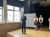 I Małopolski Wicekurator Oświaty stoi na scenie w trakcie swojego wystąpienia. Za nim stoją uczennice, a także widoczny jest napis.