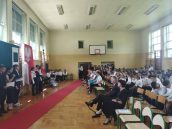 Sala gimnastyczna, gdzie odbywała się uroczystość zakończenia roku. Po prawej stronie w pierwszym rzędzie siedzi Wicekurator Oświaty, po lewej stoją uczniowie i uczennice, m.in ze sztandarem.