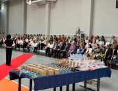 Trybuny w sali gimnastycznej gdzie odbywało się zakończenie roku, na których siedzą zaproszeni goście, w tym Małopolska Kurator Oświaty. Na bliższym planie m.in puchary i dyplomy.