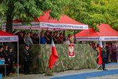 W centralnej części zdjęcia biało-czerwona flaga oraz godło, za nimi zaproszeni goście, po prawej stronie Małopolska Kurator Oświaty stoi pod dachem.