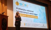 Katarzyna Gawron stoi na scenie z mikrofonem w dłoni i przemawia do publiczności w trakcie wydarzenia. Za nią wyświetlona prezentacja.