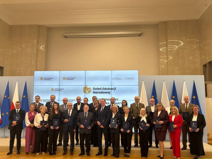 Grono honorowych Profesorów Oświaty stojących wraz z Minister Edukacji. 26 osób stojących na scenie w galowych strojach trzymających w rękach odznaczenia. W tle flagi Polski i Unii Europejskiej.