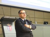 Dyrektor Szpitala Wojskowego B. Guzik przemawia do zgromadzonej publiczności do mikrofonu, ma jedną założoną rękę i wzrok skierowany na publikę.