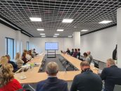 Zgromadzeni goście podczas spotkania siedzą przy stołach i obserwują prezentacje. Na stołach położone m.in notatki, woda, kawa.