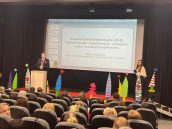 I Małopolski Wicekurator Oświaty Artur Pasek stoi na scenie przed mikrofonem i przemawia do publiczności, która siedzi w fotelach. Obok niego prowadząca wydarzenie.