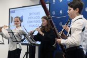 Występ artystyczny dzieci w trakcie spotkania. Uczniowie grają na instrumentach, za nimi wyświetlony baner mówiący o wydarzeniu.