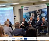 I Małopolski Wicekurator Oświaty wraz z innymi prelegentami w trakcie swojego wystąpienia, za nimi rollup, na bliższym planie publiczność.