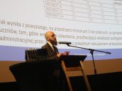 Wystąpienie prawnika w trakcie wydarzenia, za nim wyświetlona prezentacja, obok niego m.in butelki z wodą.