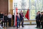 Uczniowie szkoły ubrani odświętnie, stoją przy sztandarze Szkoły, jeden z uczniów go trzyma. Reszta stoi na baczność.