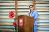 II Małopolska Wicekurator Oświaty w trakcie swojego wystąpienia, mówi do mikrofonu. Przed nią m.in czerwony kwiat. MWKO zwrócona jest do publiczności.