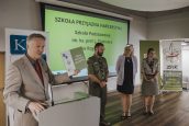 Wicekurator Artur Pasek stojący przy mównicy z tabliczką Szkoła Przyjazna Harcerstwu. W tle przedstawiciele ZHP. Za nimi banery KO oraz ZHP oraz ekran z prezentacją.