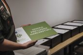 Tabliczka Szkoła Przyjazna Harcerstwu, trzymana przez kobietę w stroju harcerskim. W tle stół, a na nim wyróżnienia w ramkach.